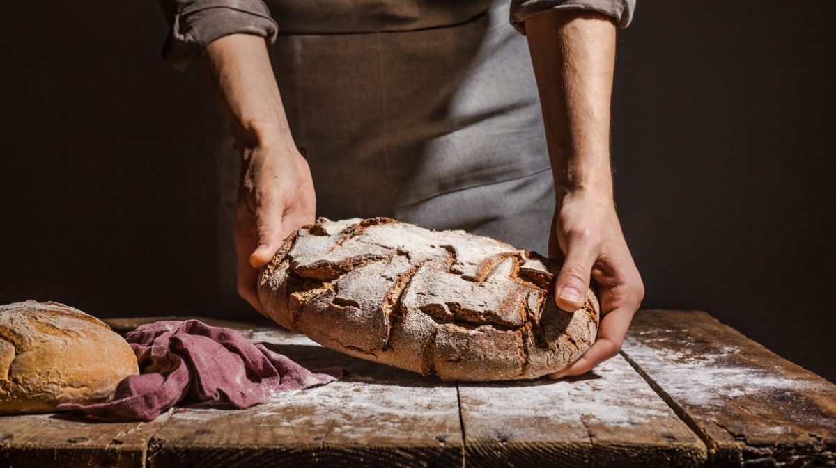 Boulanger En Tablier Manipule Une Miche De Pain Sur Une Table En Bois, Farine Et Chiffon