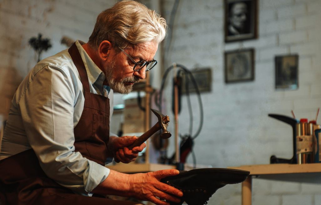 Homme Dans Son Atelier Portant Un Tablier Confectionne Une Chaussure, Il Tient Un Marteau