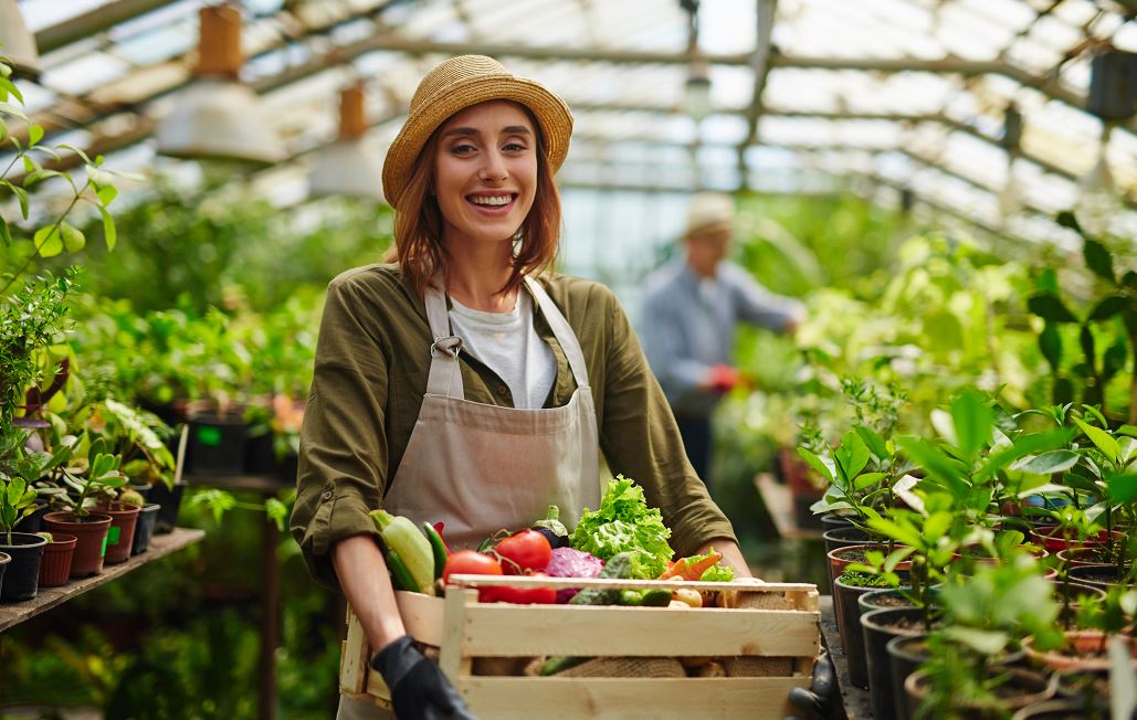 Jeune Femme Souriante Tenant Une Cagette De Légumes Frais, Elle A Mis Un Petit Chapeau De Paille Très Tendance